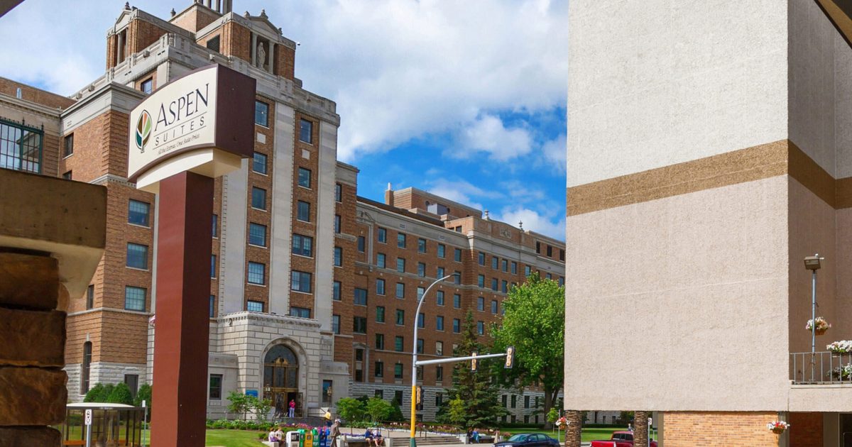 View from under an archway with stone columns, looking toward a street and the Aspen Suites hotel sign. In the background are multi-story brick and beige buildings, with a blue sky and some clouds above. Flowers are in the foreground.