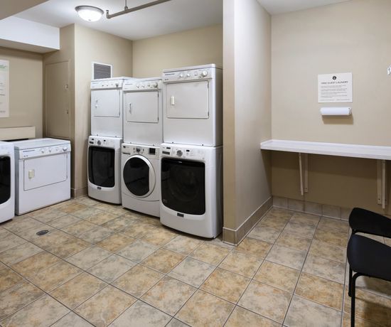 Laundry room with beige walls and tiled floor. Six white washers and dryers are stacked along the left wall. A folding table is mounted on the right wall above a black chair. Signage and safety instructions are posted on the walls.