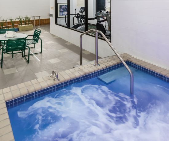 A small indoor swimming pool with bubbling water is adjacent to a fitness room visible through large glass windows. Green chairs and a table are on the tiled floor nearby, with a circular mirror and plant decorations on the wall.