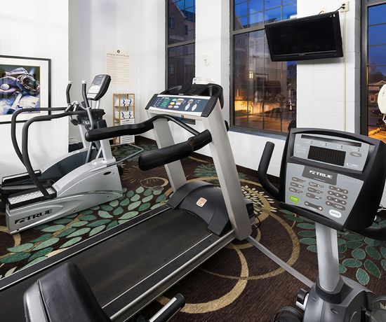 Fitness room with two treadmills and an elliptical machine on a patterned carpet. Large windows reveal a view of buildings at dawn or dusk. A wall-mounted TV is visible, along with framed sports photos.
