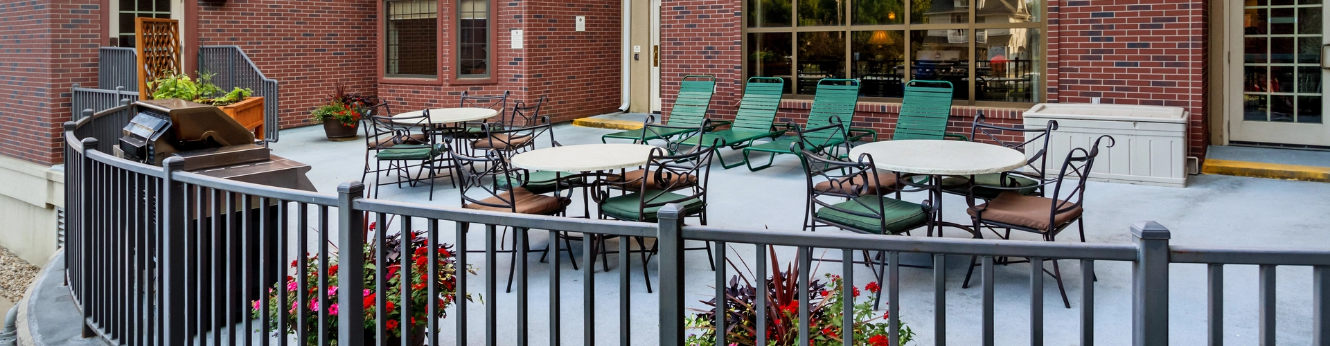 Outdoor patio area with multiple tables and chairs on a concrete surface, surrounded by a metal railing with potted flowers. Large brick building with expansive windows and a doorway in the background.