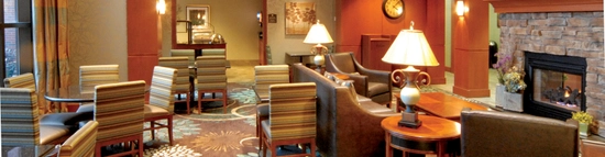 A cozy hotel lobby with patterned carpet, seating areas featuring armchairs and round tables, a stone fireplace, and a wall-mounted TV. Warm lighting and decorative elements, including plants and artwork, create an inviting atmosphere.