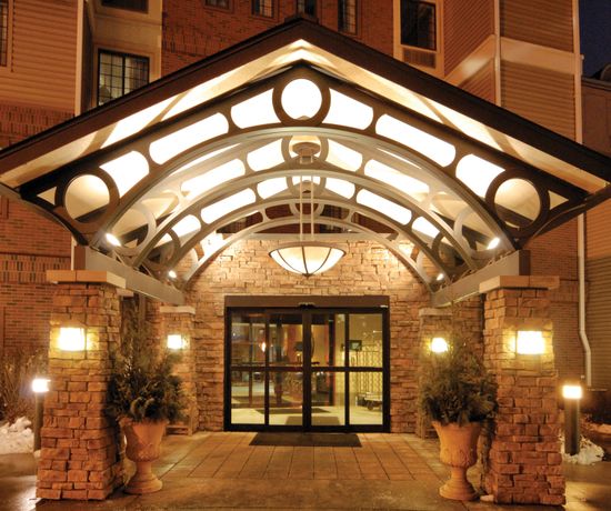 A well-lit hotel entrance with glass double doors framed by a stone archway. The structure includes detailed metalwork with circular designs. Potted plants sit on either side of the door on a tiled pathway. Snow is visible on the ground.