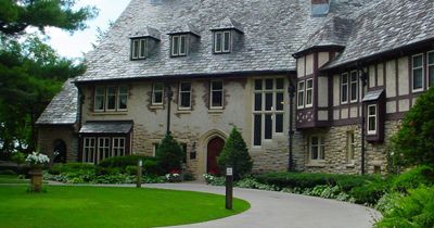 A large, historic stone building with a slate roof and numerous small windows. The entrance features an arched wooden door. A curved driveway bordered by green lawns and shrubs leads to the entrance.