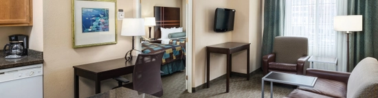 Hotel room with a living area featuring a sofa, armchair, coffee table, and TV. A desk and chair sit near a kitchenette with a counter and coffee maker. A window with curtains is in the corner. A bedroom is visible through an open door.