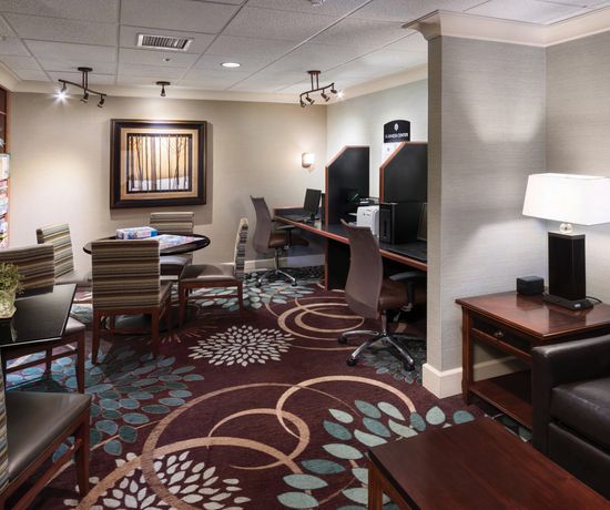 A cozy hotel business center featuring three computer stations with chairs, a small round table with four chairs, decorative plants, patterned carpet, and a lamp on a side table. A framed artwork hangs on the wall.