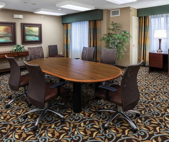A conference room with a large oval wooden table surrounded by brown chairs on a patterned carpet. The room is decorated with paintings, plants, and a lamp by the window, providing a warm and professional atmosphere.